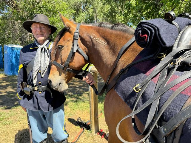 An individual in period gear poses next to a brown horse