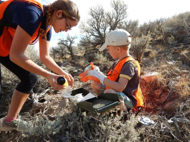 A teenager helps a child open a box on a mountainside.