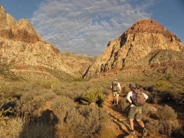 Two hikers in shorts walk through a dramatic red and brown cliffs.