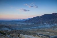 Wind turbines lined across a landscape with scattered vegetation and mountains in the background. 