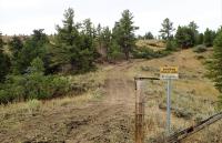 ALT-TEXT – Photo. Foreground: a yellow-colored “Entering Public Land” sign mounted on a grey-colored, metal post is positioned near the gate opening in a fence line comprised of round wooden posts and metal wire. Background: a scenic view of the Bullwhacker area of the Upper Missouri River Breaks National Monument; a dirt road curves slightly uphill through yellow and green grasses dotted with sagebrush; conifer trees and bushes grow in scattered clumps on the hills; overcast, grey-colored sky.