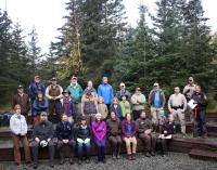 A diverse group of people gather for an outdoor photo in front of spruce trees. They are dressed for volunteer trail work.