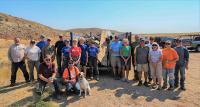 A group of about 20 people and a dog pose for a group photo in front of a large dumpster filled with garbage.