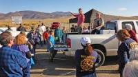 A large group of people standing in the desert listening to someone speaking from the back of an open bed truck.