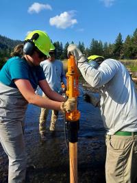 Addison Schuette drives wooden post into the streambed using a hydraulic post-pounder with help from Casey Schuder and Gabriel Juarez