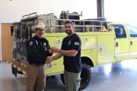 two people shake hands in front of a yellow fire engine. One person hands keys to the other.