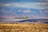 A small airplane sprays herbicide onto grasses. Canyons are in the background.