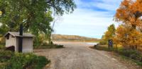 Photo. Daytime; partly cloudy blue skies. Gravel road entrance to a riverside developed recreation area. Vault toilet building on the left side of the frame; visitor kiosk sign on the right side of the frame. Trees and bushes beginning to change to autumn colors; river and rocky, cliff-like far riverbank in the background. 
