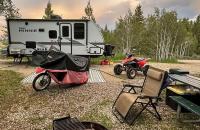 A campsite in the woods contains a travel trailer, covered motorcycle, ATV, picnic table, camping chair and a fire grate.