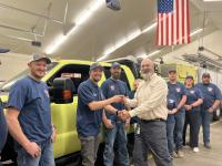 An image of men and women in jeans an blue tee shirts lined up in front of a yellow-green wildland fire engine inside the volunteer fire department warehouse. A man wearing a taupe BLM dress shirt smiles and shakes hands with a smiling man in jeans and a blue tee shirt who accepts the handshake and keys to the fire engine. 