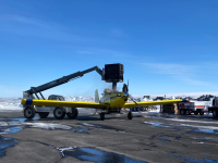A yellow airplane on tarmac with a snowy backdrop, a large crane holding a box overhead
