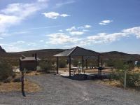 A photo showing a gravel parking spot next to a covered picnic table. A concrete path leads to a double-vault toilet, several other campsites with covered picnic tables can be seen in the background.  