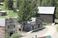A photo showing several historic wooden buildings with a dirt road going through the town. Wood ramps lead to the entrance of each building. Two large pine trees flank the central building. 