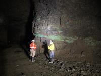 Two miners wearing safety gear inspect a rocky underground wall with green mineral deposits illuminated by a flashlight.