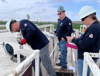 Three workers wearing hard hats and navy shirts conduct an inspection on industrial equipment outdoors with green fields in the background.