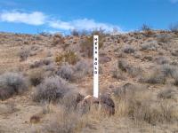 A white wooden claim marker stands upright in a desert landscape, surrounded by dry grasses, rocks, and scattered shrubs.