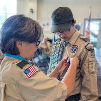 A Boy Scout in uniform looks down as an adult leader pins the Eagle Scout medal to his chest. The scout wears a sash covered in merit badges, and other scouts are visible seated in the background.
