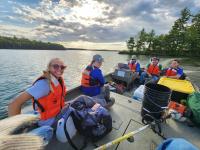 A group of individuals wearing orange life jackets sits in a boat on a lake, surrounded by trees and a cloudy sky.
