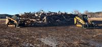 Two yellow skid steer loaders parked on either side of a large pile of cut tree logs and branches in an open field under a clear blue sky.