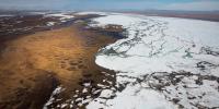 A landscape showing a frozen body of water meeting brown land is seen from a high elevation, with a blue sky above.