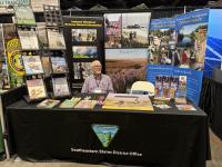 Person sitting at an expo booth surrounded by pamphlets and informational displays