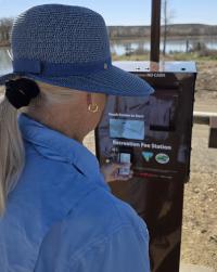 Woman in a blue hat and jacket using a touchscreen recreation fee station outdoors near a river.