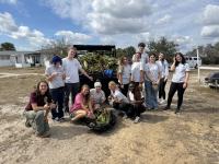 Group of young volunteers posing outdoors with collected plants and yard waste on a sunny day.