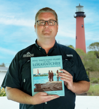 Person holding a book titled "Five Thousand Years on the Loxahatchee" with a lighthouse and trees in the background.