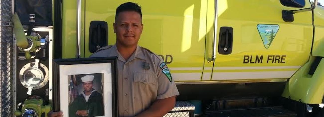 BLM employee standing in front of BLM fire truck holding photo of himself in military uniform.