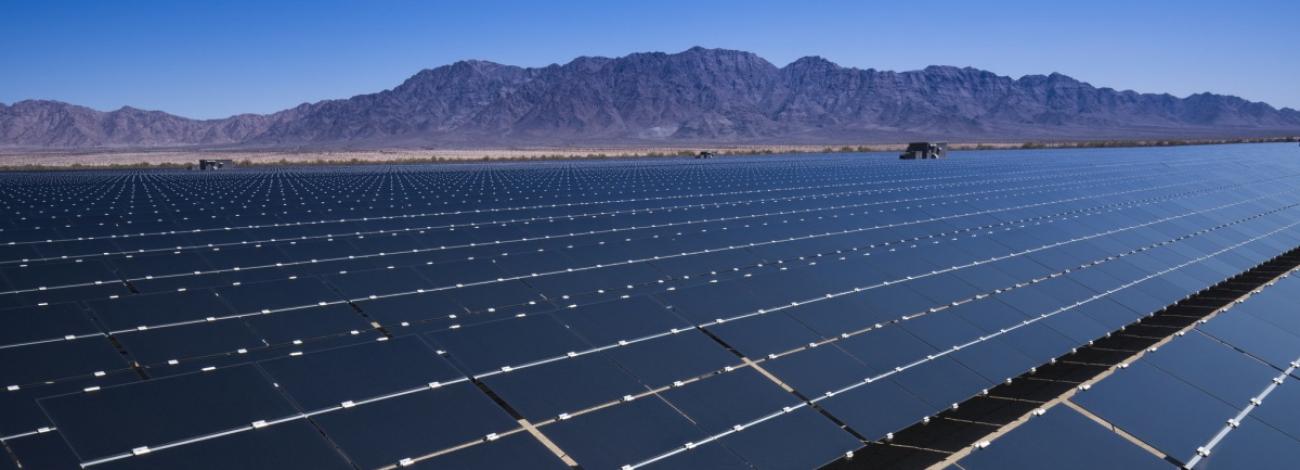 Image shows numerous rows of solar panels in the middle of the California desert field surrounded by mountains under a clear sky.