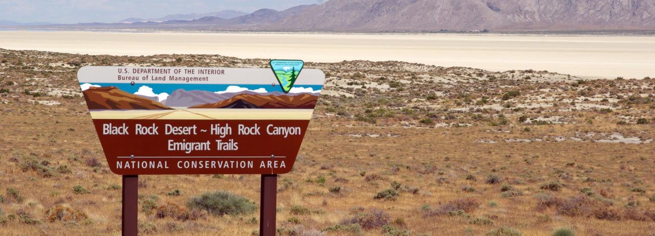 Entrance sign among grasses in front of a desert playa and mountains