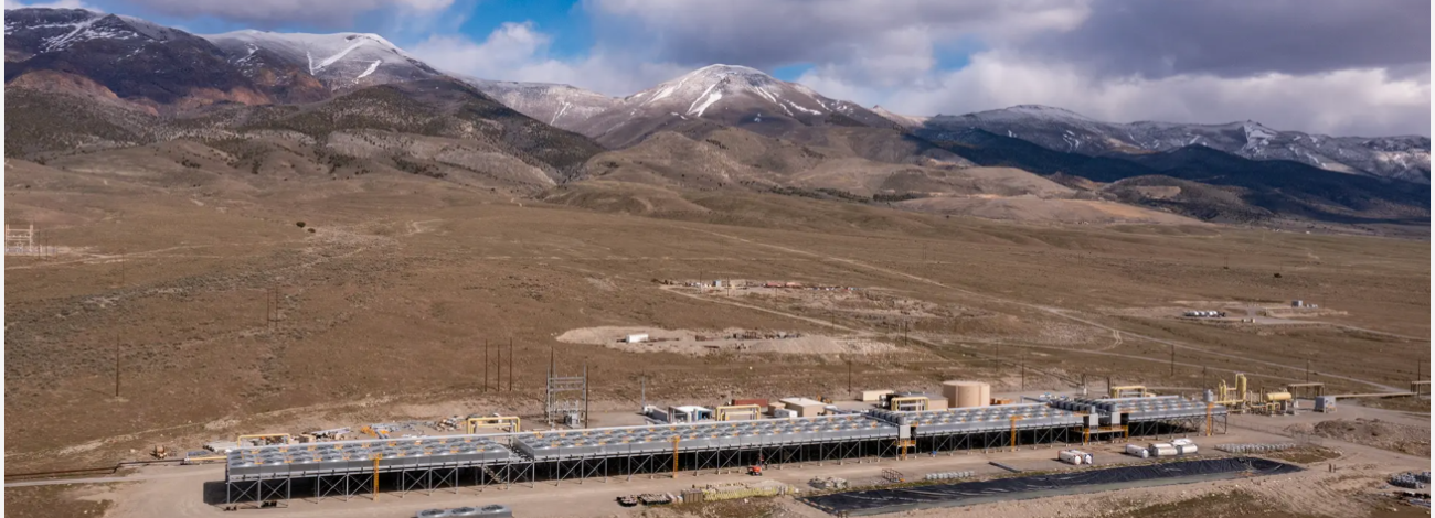 A landscape scene with brown hills and a partly cloudy sky. The foreground is taken up by a long black low-slung structure, a geothermal power plant.