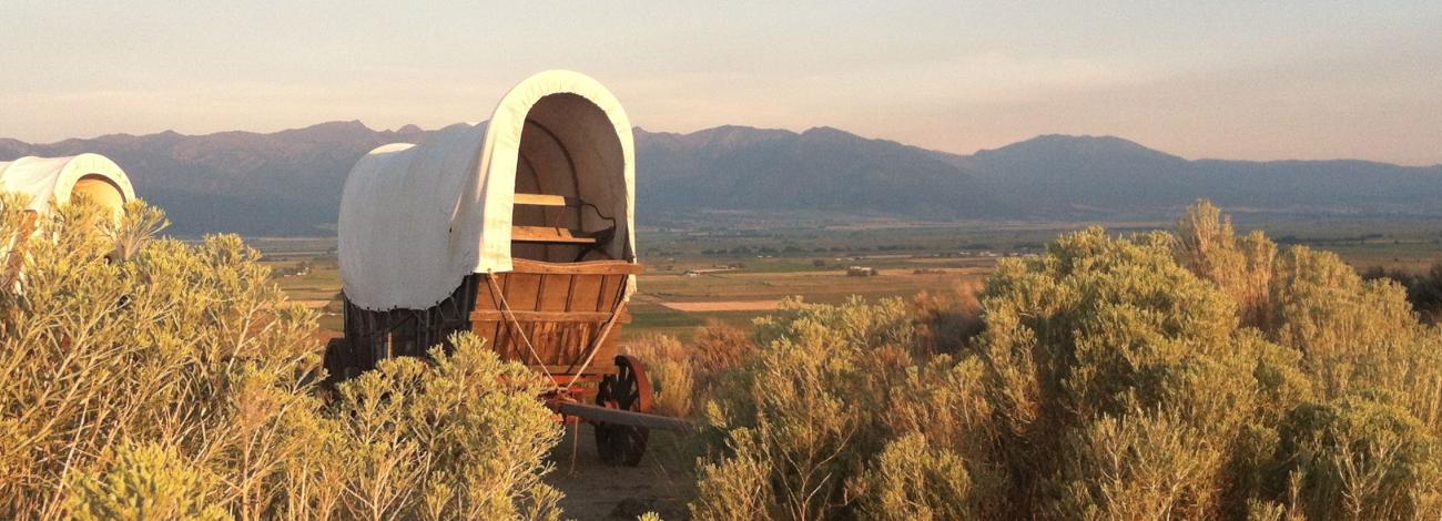 A covered wagon surrounded by sage brush with distant mountains at sunset