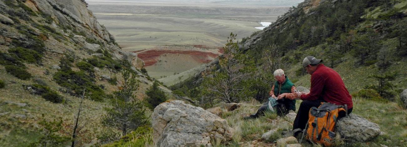 Two hikers with back packs on top of a large hill overlooking a big valley.