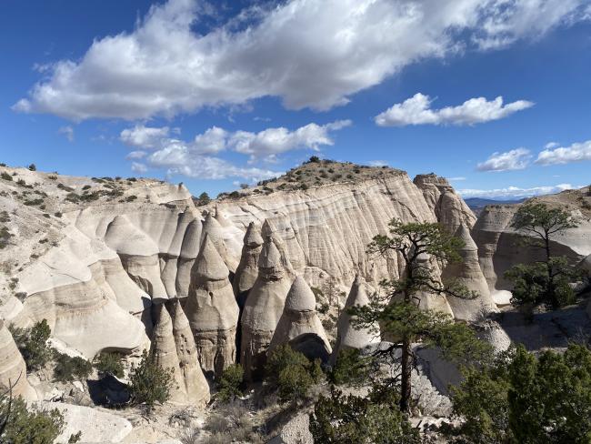 Eroded rock formations and cone-shaped hoodoos rise from a desert landscape under a partly cloudy blue sky, with sparse trees in the foreground.