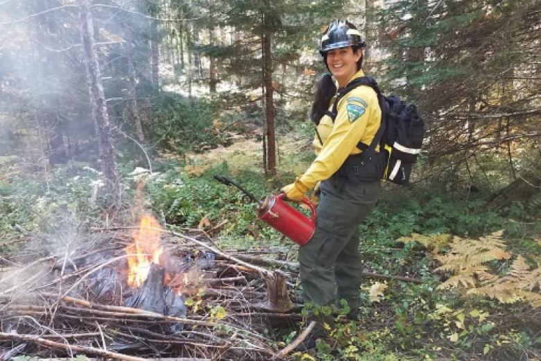smiling firefighter in traditional yellow shirt holding a drip torch near a burn pile