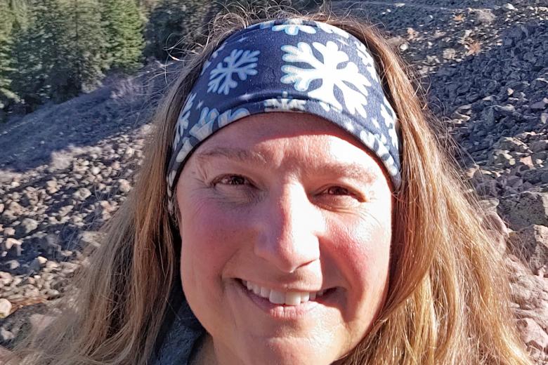 Photo. Foreground: head-and-shoulders view of a woman, smiling, with reddish-brown, long hair, fair-skinned, wearing a snowflake-printed headband and blue zip-up jacket. Background: outdoors, daytime, sunny, forest and rocky hillside.