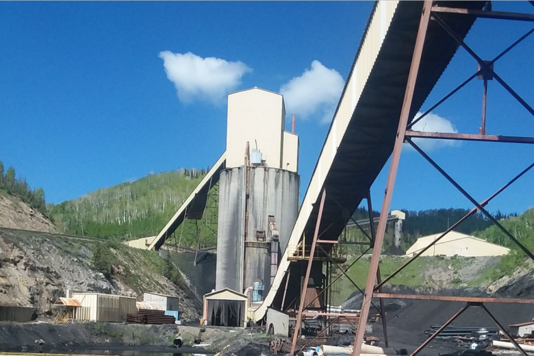 Industrial mining facility with tall silos and conveyor belts set against green hills under a clear blue sky.