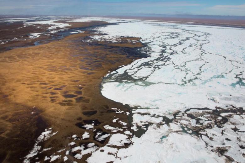 A landscape showing a frozen body of water meeting brown land is seen from a high elevation, with a blue sky above.