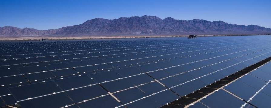 Image shows numerous rows of solar panels in the middle of the California desert field surrounded by mountains under a clear sky.