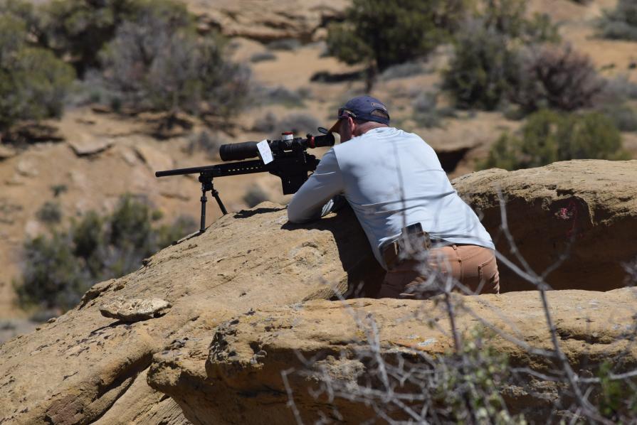 A photo of a man leaning on a rock in the desert, aiming a firearm into the distance.