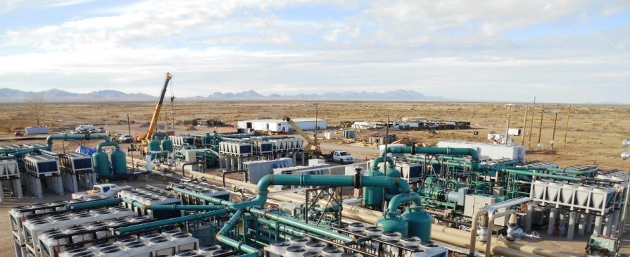 The image shows a large geothermal plant in an open field of dead grass under a cloudy sky.