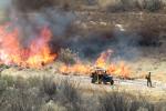 a fire burns through vegetation as firefighters look on