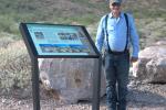 Person wearing suspenders and a ball cap standing next to an interpretive panel