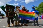 Four mascots holding a pride flag with blue sky and white clouds above.