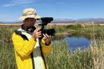 The image shows a man wearing a brimmed hat and yellow jacket standing near a wetland area with mountains in the background. He is looking down into the viewfinder of his video camera with headphones around his neck. 