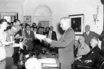 Image shows President Harry S. Truman standing at a desk and announcing the surrender of Japan to a group of reporters.