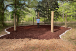 A teenaged boy stands in between four large posts in a hammock area he created with wooded area seen behind him. 