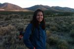 Vanessa Lacayo, smiling and wearing a blue jacket, stands in a field of sagebrush. Behind her is a line of small mountains, brownish green in color.