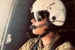 A Caucasian person in a white helmet with a black visor and mic attached and a brown flight suit sits inside an aircraft looking out the window.